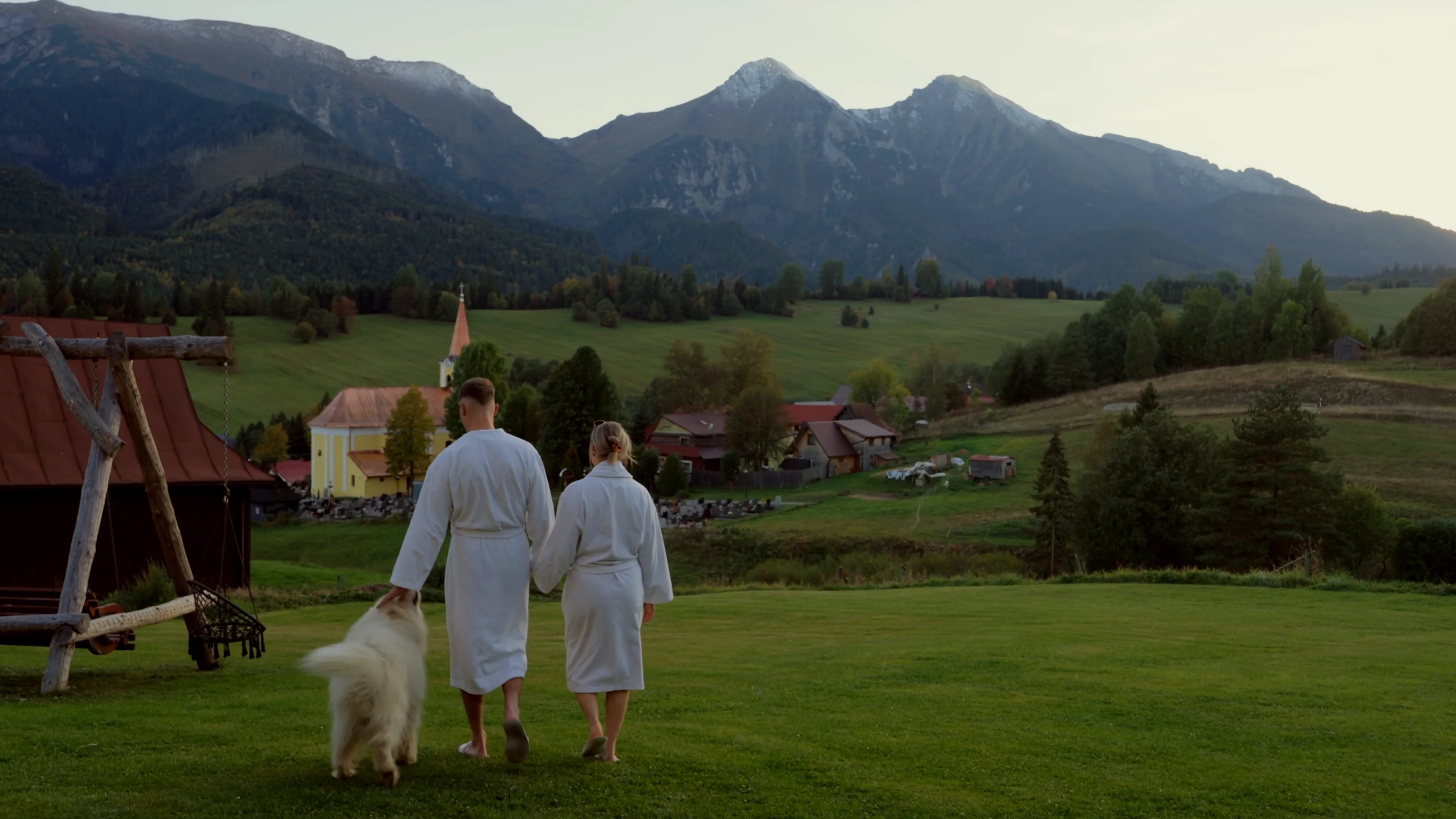 A couple in bathrobes walking across a meadow with a Samoyed, with a swing and the Belianske Tatras behind them