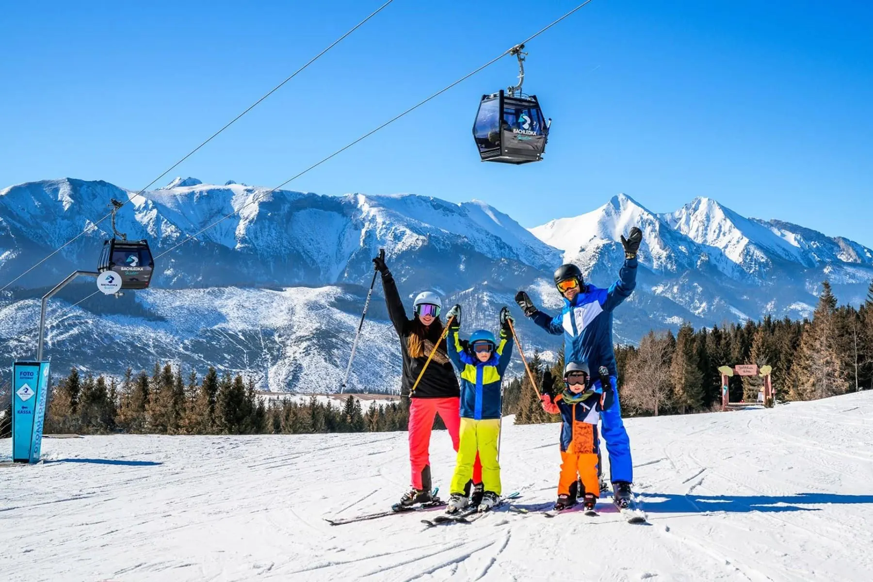 Family on skis beneath the gondola at Bachledka, with the snow-covered Belianske Tatras behind