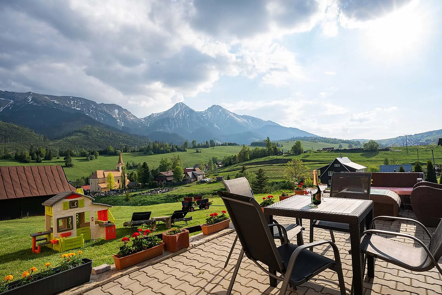 Terasa so stolom a stoličkami, pod ňou lúka s lehátkami a záhradným domčekom, v pozadí kostol a Belianske Tatry